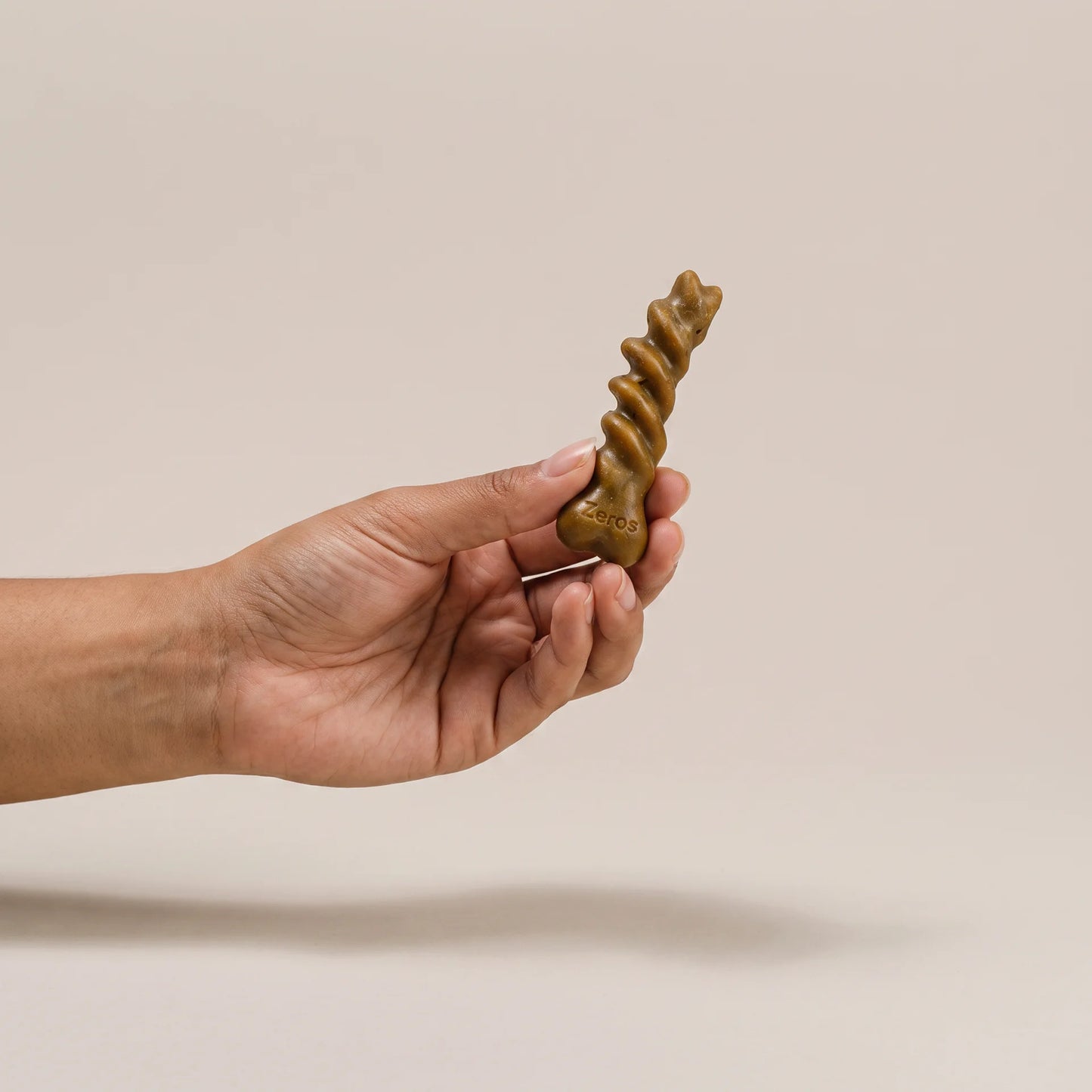 A hand holding a small brown, spiral-shaped dental chew against a plain beige background.
